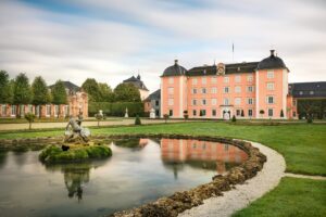 Schloss mit Garten und Teich, umgeben von Bäumen.