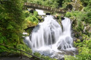 Wasserfall mit fließendem Wasser und Holzbrücke im Hintergrund, umgeben von grüner Vegetation.