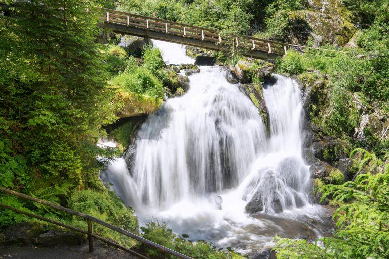 Wasserfall mit fließendem Wasser und Holzbrücke im Hintergrund, umgeben von grüner Vegetation.