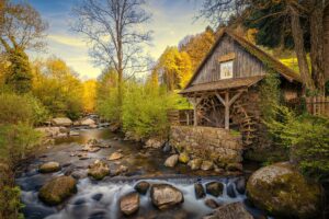 Idyllische Mühle am Fluss mit steiniger Uferlandschaft und Bäumen im Hintergrund.