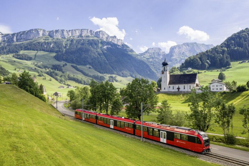 Roter Zug fährt durch eine grüne Landschaft mit Bergen und einer Kirche.