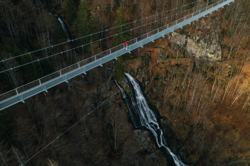 Person auf einer Hängebrücke über einen Wasserfall in einer bewaldeten Umgebung.