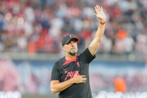 Mann mit grauem Bart und Baseballcap winkt in einem Stadion.