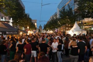 Belebte Straße mit Menschenmenge und Ständen bei Abendveranstaltung.