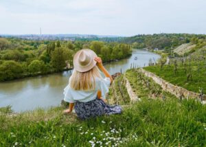 Frau mit Hut sitzt am Flussufer und genießt die Aussicht auf die Landschaft.