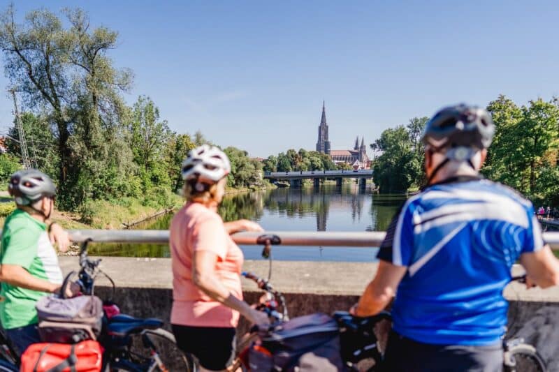 Drei Radfahrer auf einer Brücke mit Blick auf einen Fluss und eine Kirche im Hintergrund.