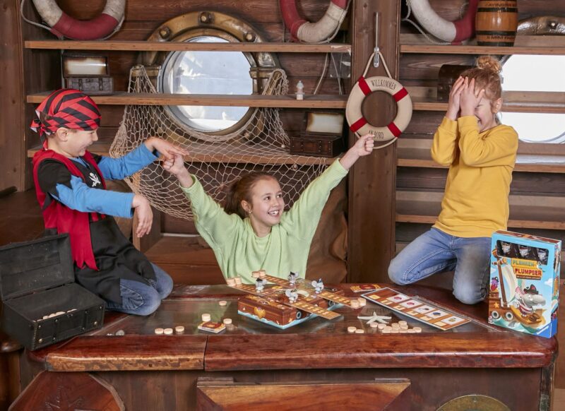 Drei Kinder spielen ein Brettspiel auf einem Schiff, zwei jubeln, während eines die Hände vor das Gesicht hält.