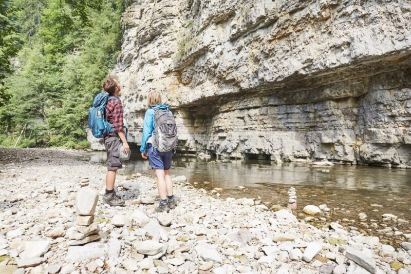 Zwei Wanderer betrachten eine Felswand an einem Flussufer.