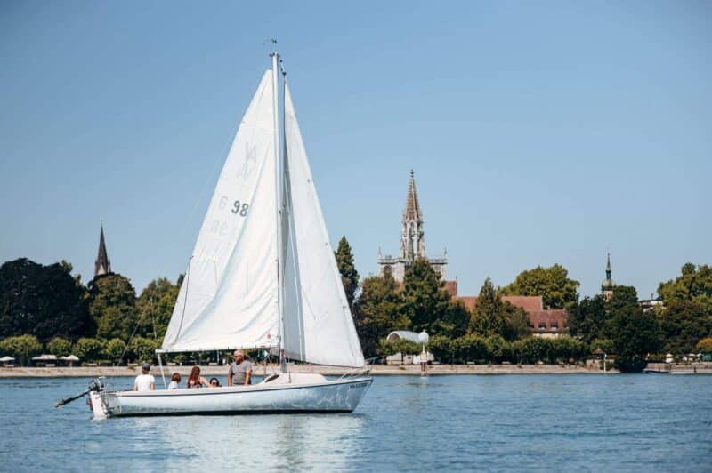 Segelboot auf ruhigem Wasser mit Bäumen und Gebäuden im Hintergrund.