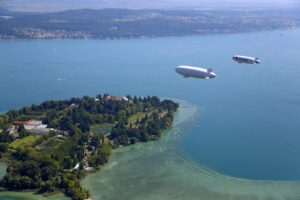 Zwei Luftschiffe über einem See mit einer bewaldeten Insel im Hintergrund.