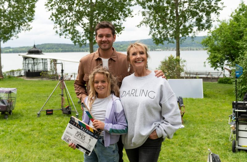 Drei Personen stehen im Freien am Wasser, eine hält ein Filmschild.