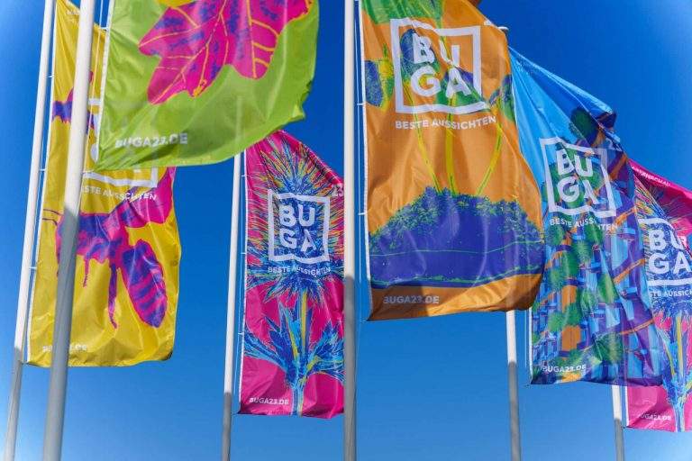 Bunte Fahnen mit dem Schriftzug 'BUGA' wehen vor blauem Himmel.