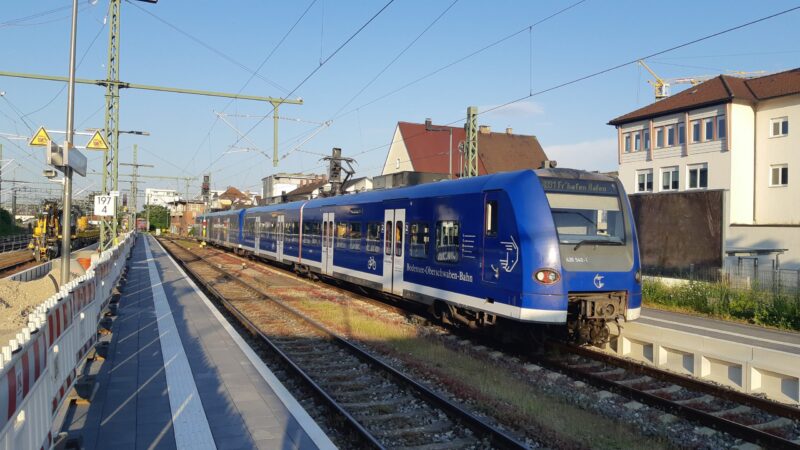 Blauer Zug an einem Bahnhof mit Wohngebäuden im Hintergrund.