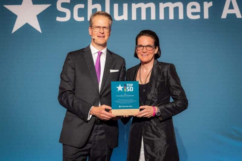 Zwei Personen halten eine Auszeichnung auf einer Bühne mit blauem Hintergrund.