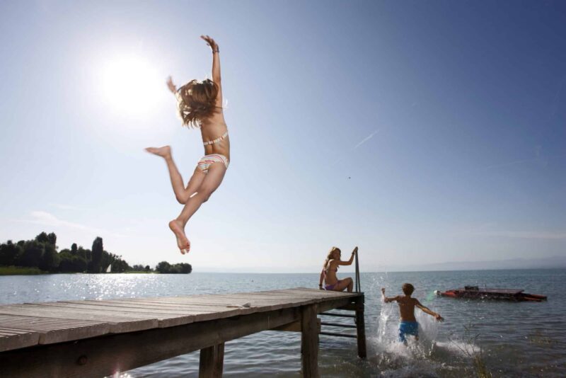 Mädchen springt vom Steg ins Wasser, zwei Kinder spielen am Ufer.