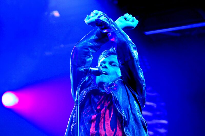 Mann mit Mikrofon in Lederjacke, hebt die Arme während eines Auftritts.