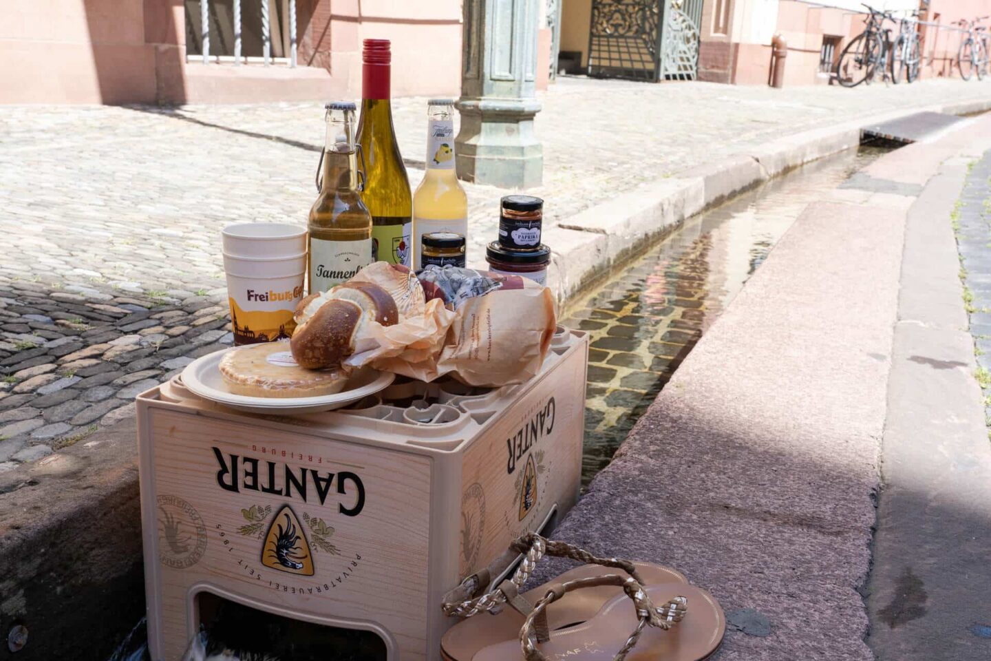 Holzkiste mit Getränken und Lebensmitteln an einem gepflasterten Weg.