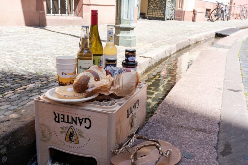 Holzkiste mit Getränken und Lebensmitteln an einem gepflasterten Weg.