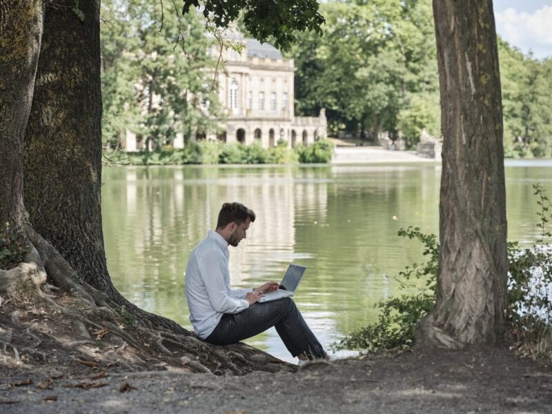 Mann sitzt am Ufer eines Sees und arbeitet am Laptop.
