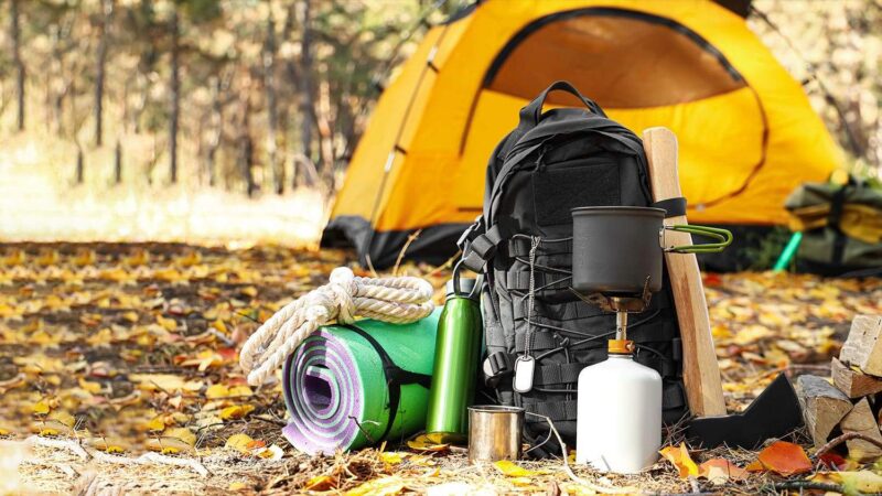 Campingausrüstung mit Rucksack, Kocher und Schlafmatte vor einem Zelt.
