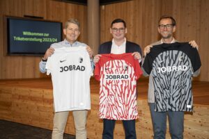Drei Männer präsentieren neue Fußballtrikots in einem modernen Raum.