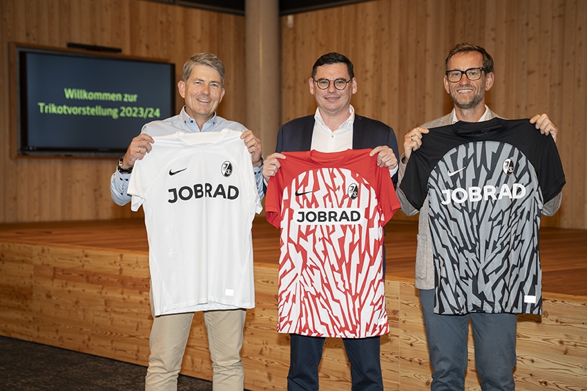 Drei Männer präsentieren neue Fußballtrikots in einem modernen Raum.