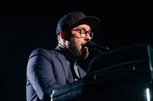 Mann mit Brille und Hut singt am Mikrofon, spielt Klavier.