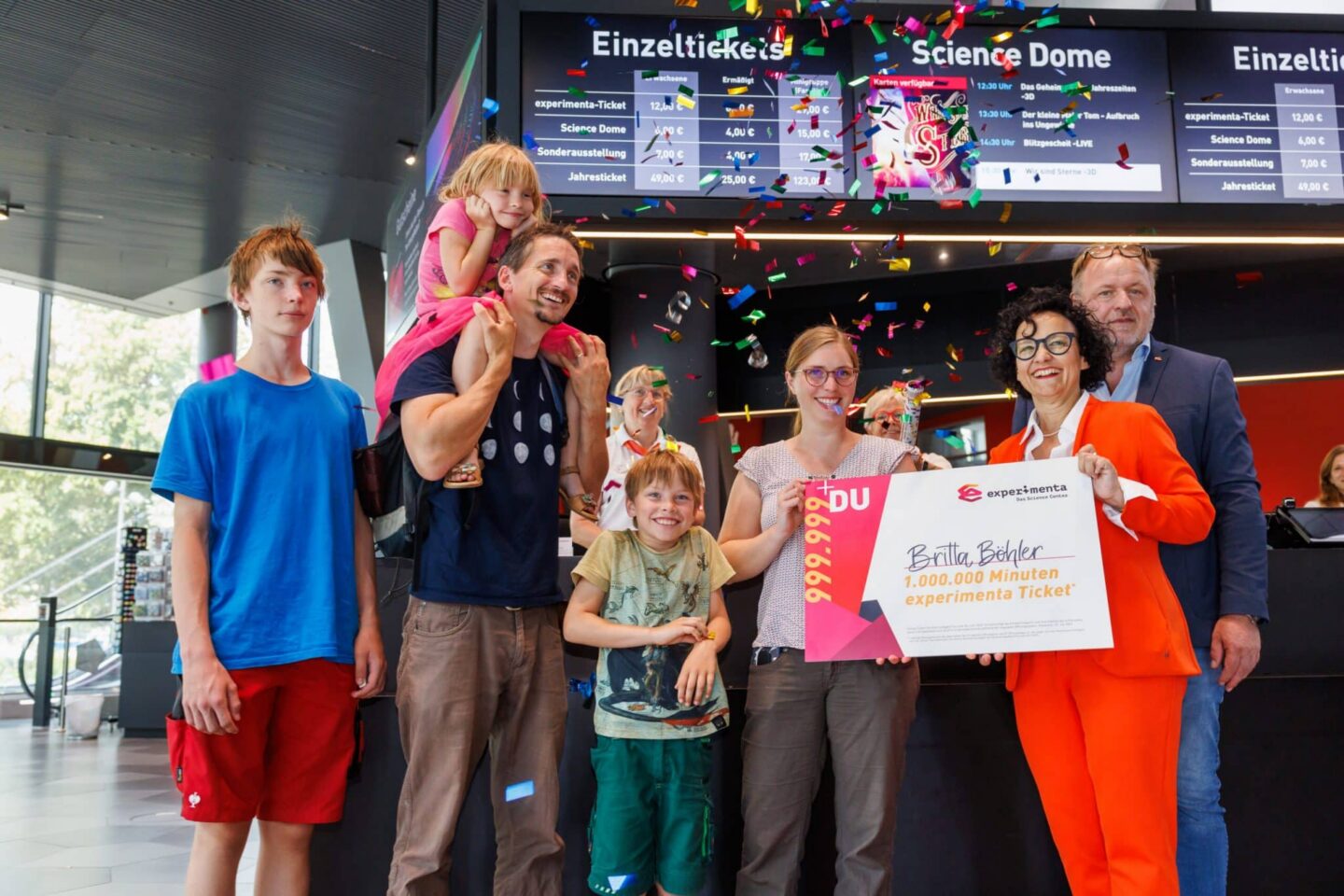 Familie feiert mit Konfetti und einem großen Ticket in einem modernen Gebäude.