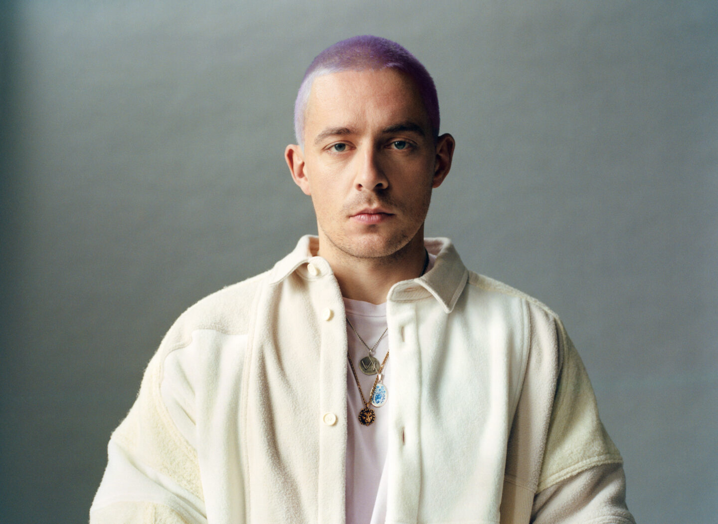 Mann mit lila Haaren und Schmuck, trägt eine weiße Jacke vor neutralem Hintergrund.