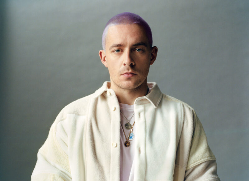 Mann mit lila Haaren und Schmuck, trägt eine weiße Jacke vor neutralem Hintergrund.