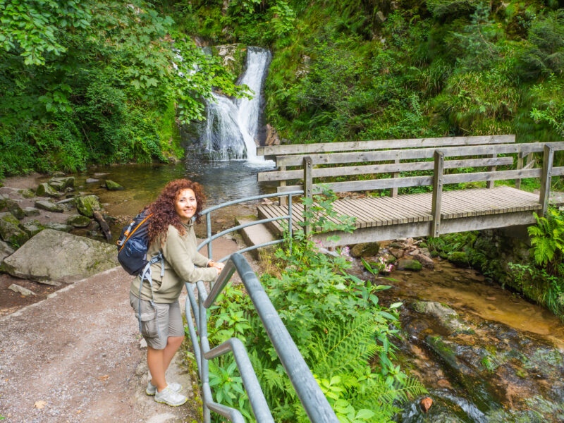 Frau mit Rucksack steht an einem Wasserfall in der Natur.