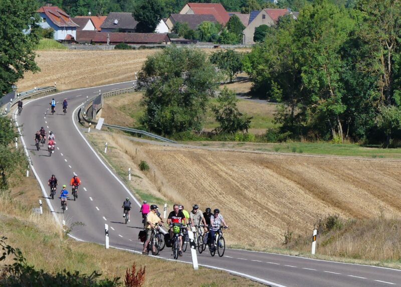 Gruppe von Radfahrern auf einer kurvenreichen Landstraße in ländlicher Umgebung.