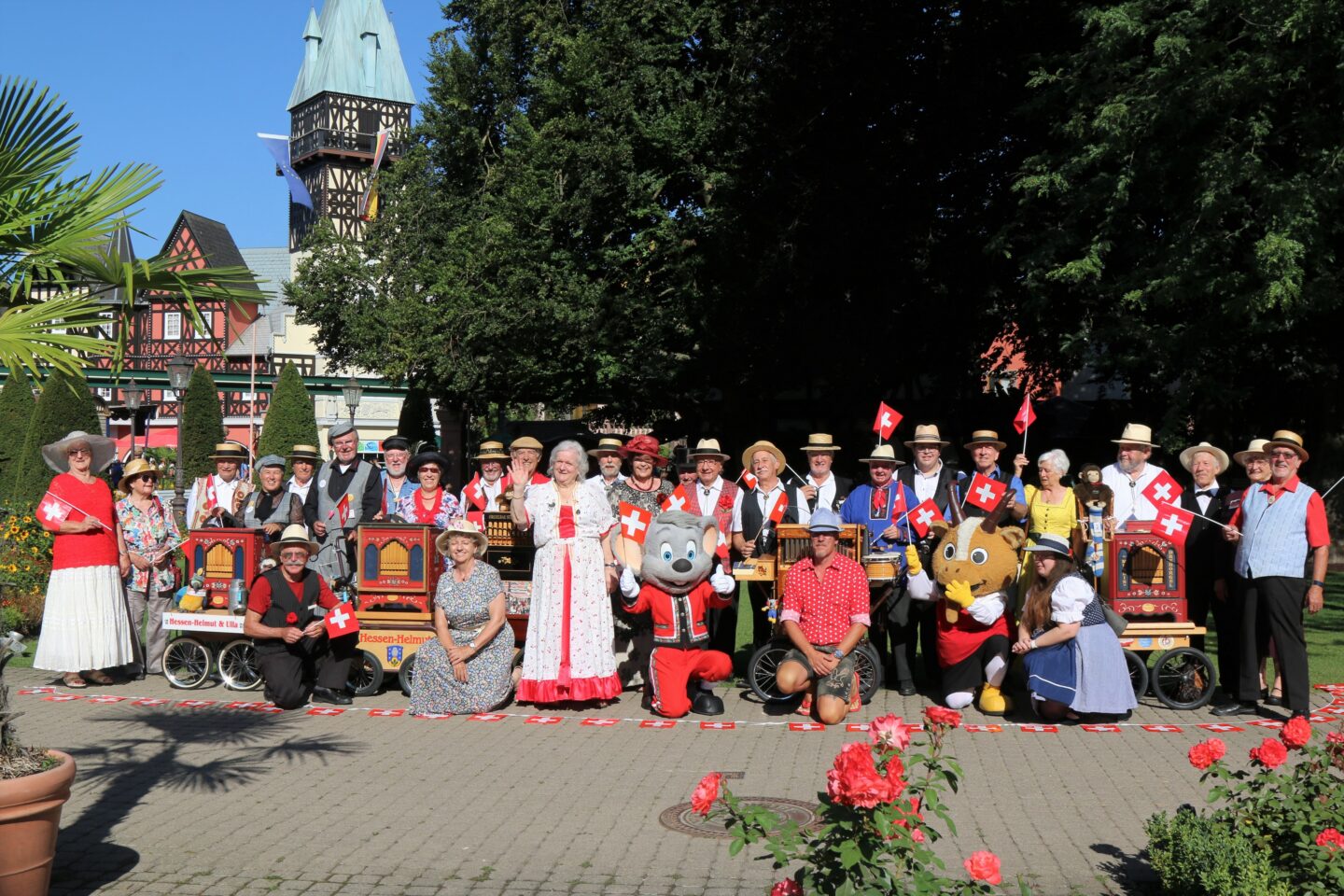 Gruppenszene mit Menschen in traditioneller Kleidung und Maskottchen, umgeben von Blumen.