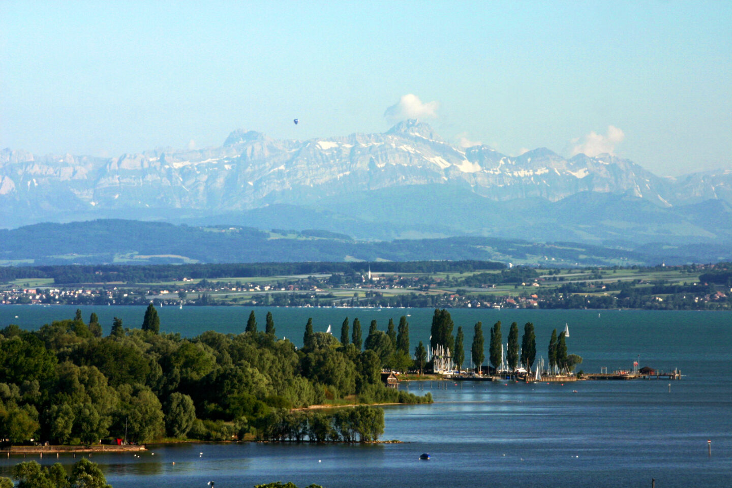 Panorama mit See, Bäumen und schneebedeckten Bergen im Hintergrund.