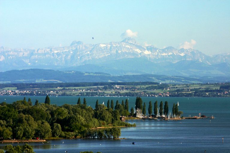 Panorama mit See, Bäumen und schneebedeckten Bergen im Hintergrund.