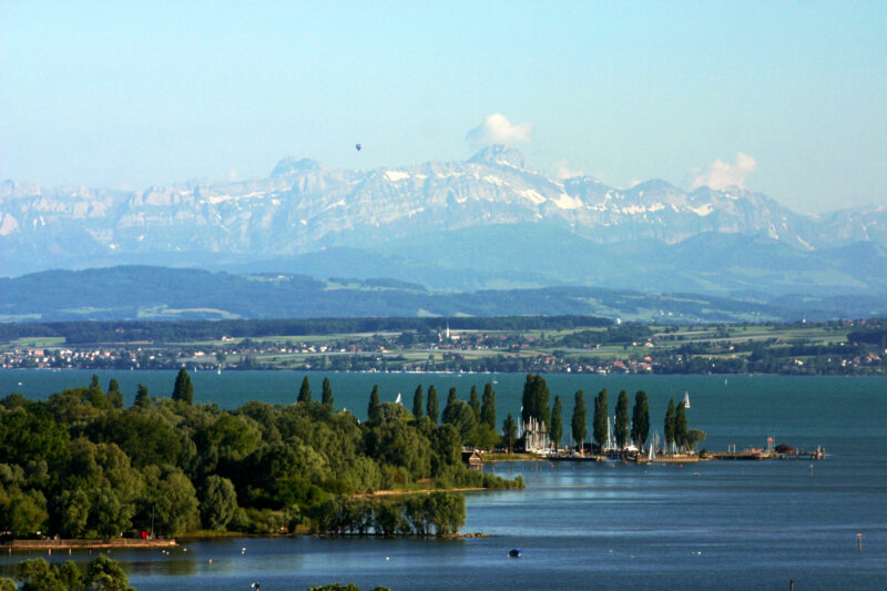 Panorama mit See, Bäumen und schneebedeckten Bergen im Hintergrund.