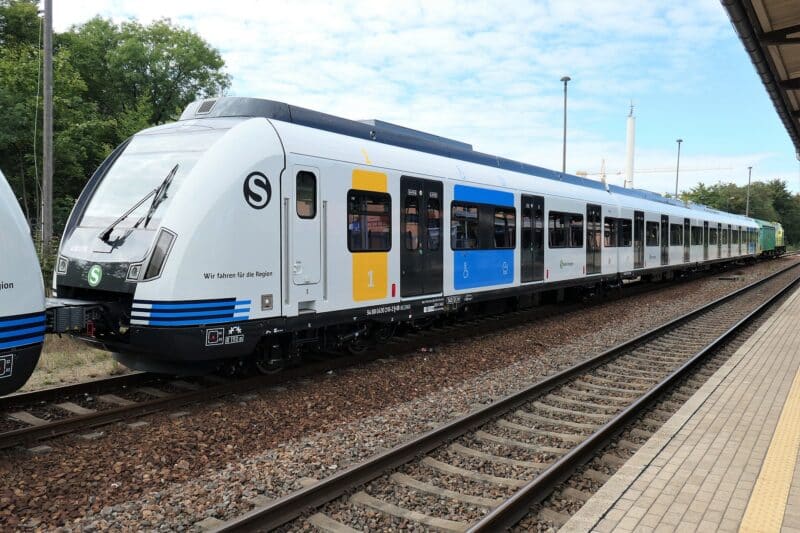 Moderner S-Bahn-Zug an einem Bahnhof mit Gleisen und Bäumen im Hintergrund.