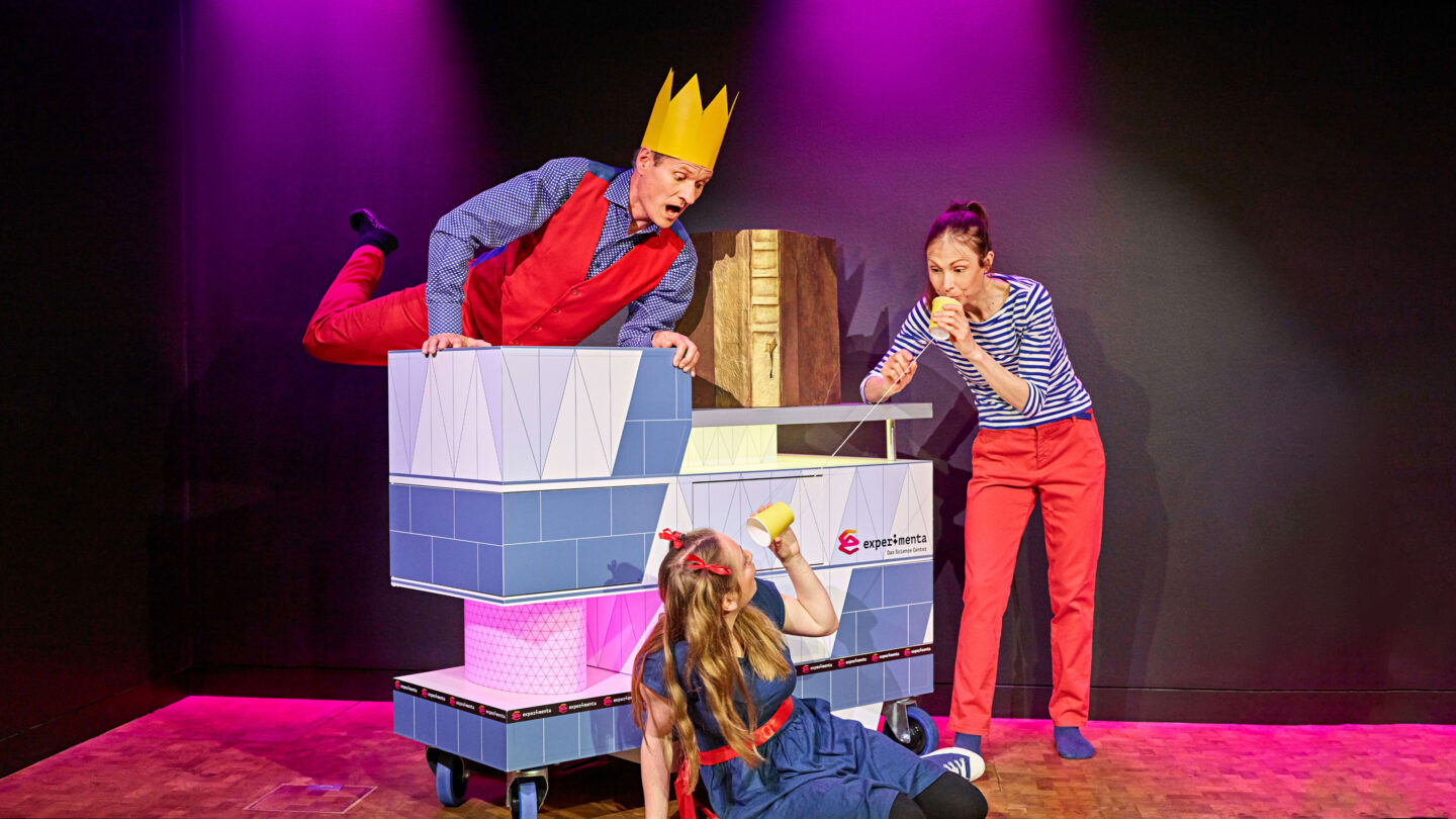 Drei Schauspieler in bunten Kostümen auf einer Bühne mit lila Beleuchtung.