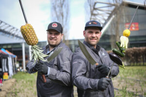 Zwei Männer mit großen Messern und Obst/Gemüse im Freien.
