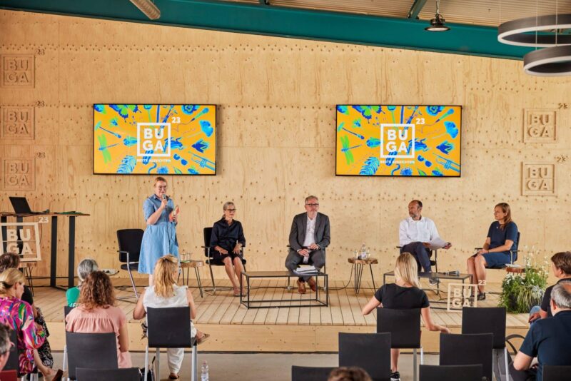 Podiumsdiskussion mit mehreren Personen und Bildschirmen im Hintergrund.