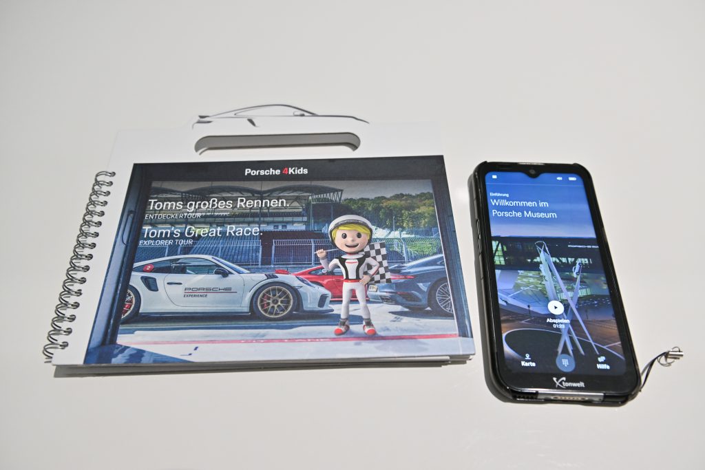 Das Porsche 4Kids Entdeckerbuch für die Tour mit Tom Targa (links) und der Porsche 4Kids Multimedia Guide. Foto: Porsche AG - Stuttgart