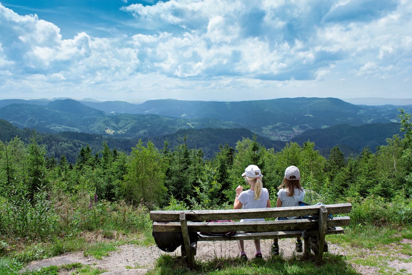 Zwei Mädchen sitzen auf einer Bank mit Blick auf eine grüne Berglandschaft.