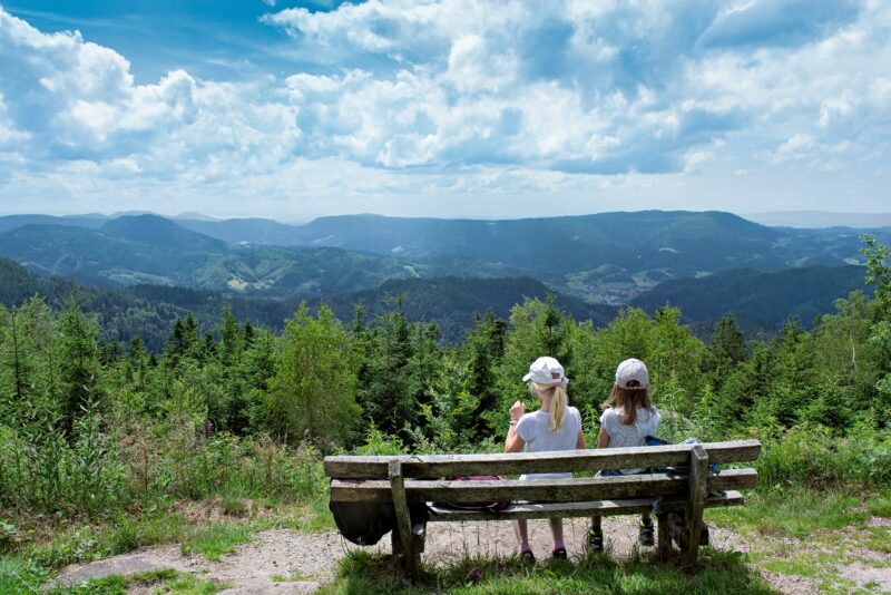 Zwei Mädchen sitzen auf einer Bank mit Blick auf eine grüne Berglandschaft.
