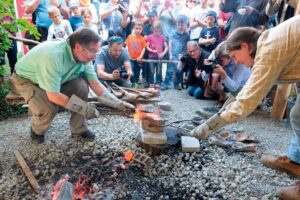 Zwei Männer schmieden mit Feuer, umgeben von einer Gruppe von Zuschauern.