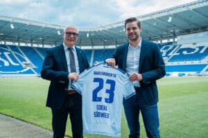 Zwei Männer halten ein Fußballtrikot des Karlsruher SC in einem Stadion.