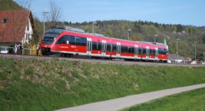 Roter Zug fährt entlang einer grünen Wiese mit Bäumen im Hintergrund.