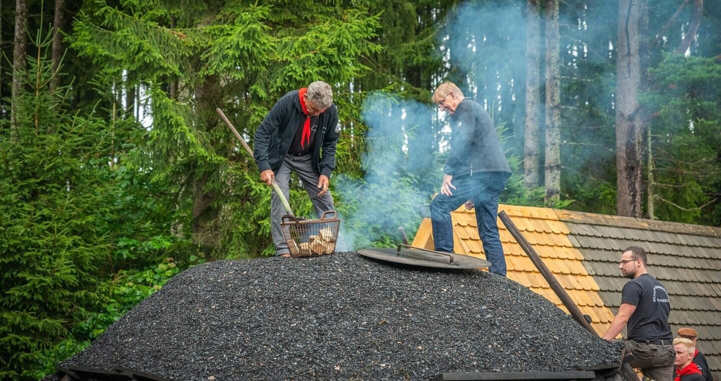 Männer arbeiten auf einem Dach, Rauch steigt auf, Wald im Hintergrund.