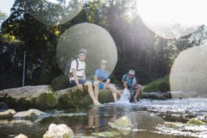 Drei Personen sitzen am Ufer eines Flusses und genießen die Sonne.