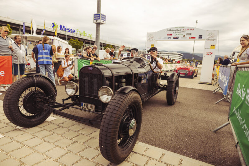 Oldtimer-Rennwagen beim Start eines Events mit Zuschauern und Banner im Hintergrund.