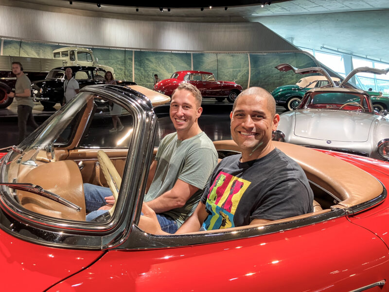 Zwei Männer sitzen in einem roten Oldtimer in einem Automuseum.